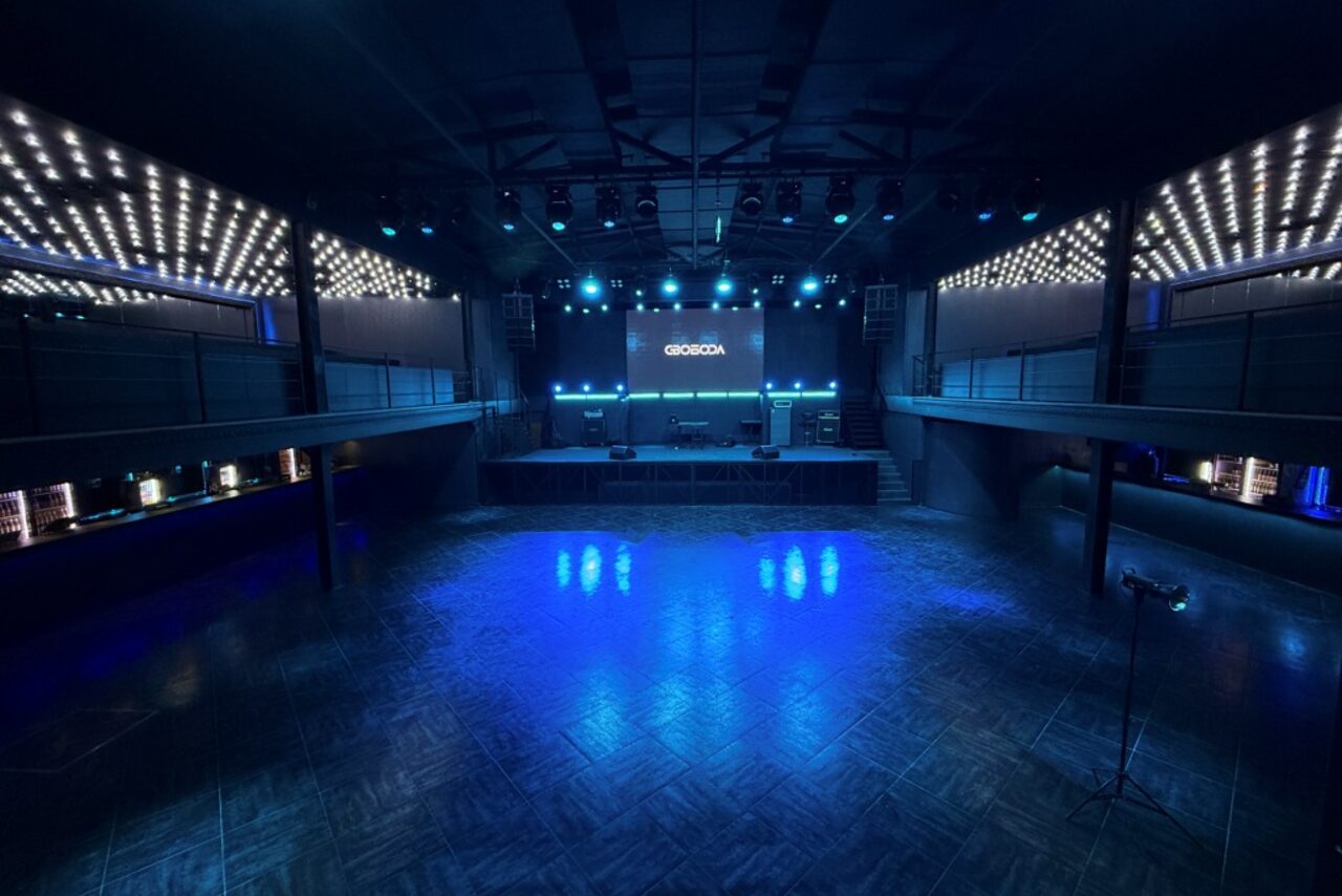 Арендовать СВОБОДА. Зал на 1200 человек. Большое сцена с синим освещением, пустым танцполом перед ней, окружена балконами по периметру. Панельные стены и потолок, на сцене оборудование, светодиодные панели и светильники по краям.