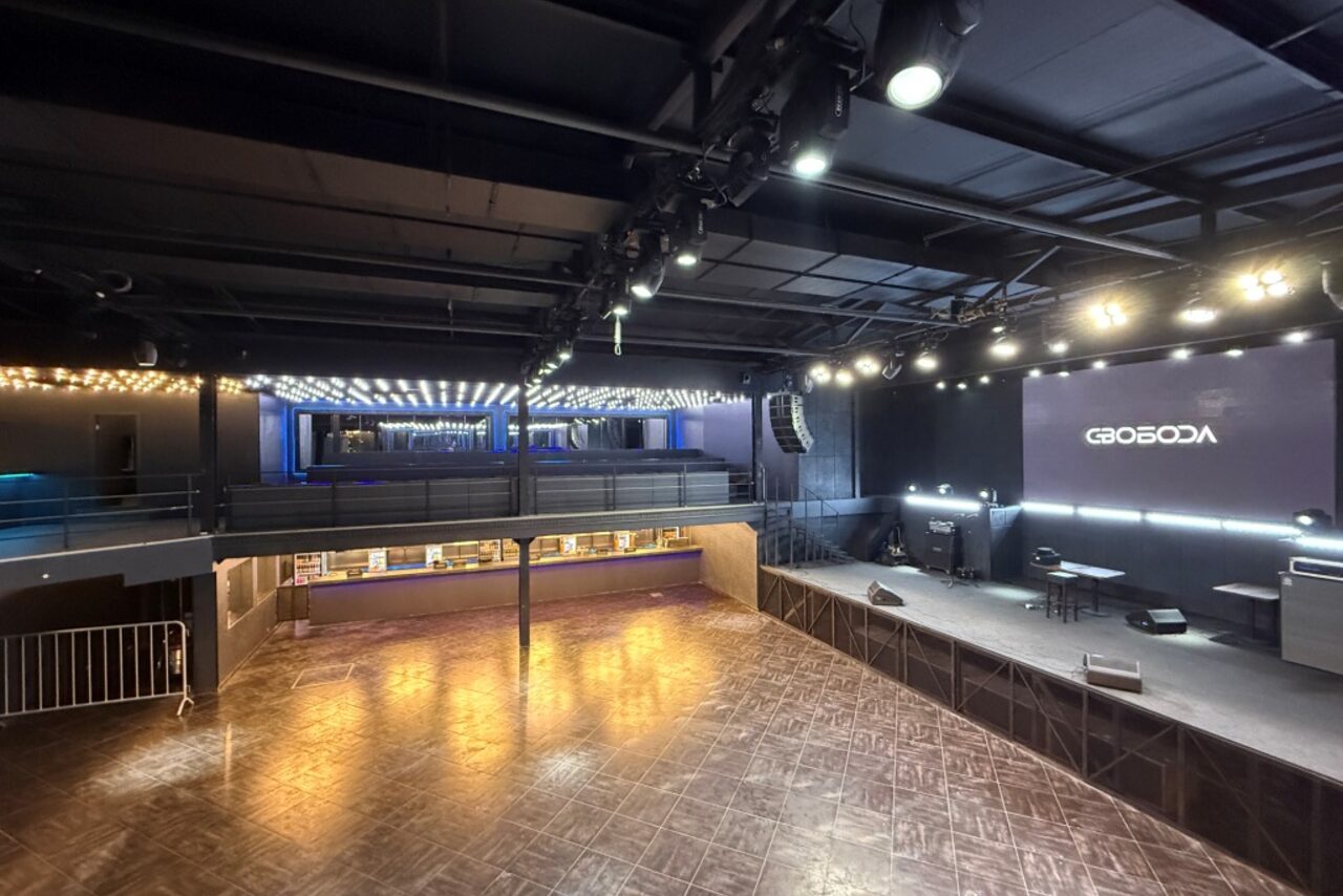 Арендовать СВОБОДА. Зал на 1200 человек. Просторное помещение с высокой сценой, деревянным полом и металлическими перилами. На сцене стоят столы и стулья. Есть светодиодное освещение и большой экран. Черный потолок с многочисленными светильниками. На экране надпись на кириллице.