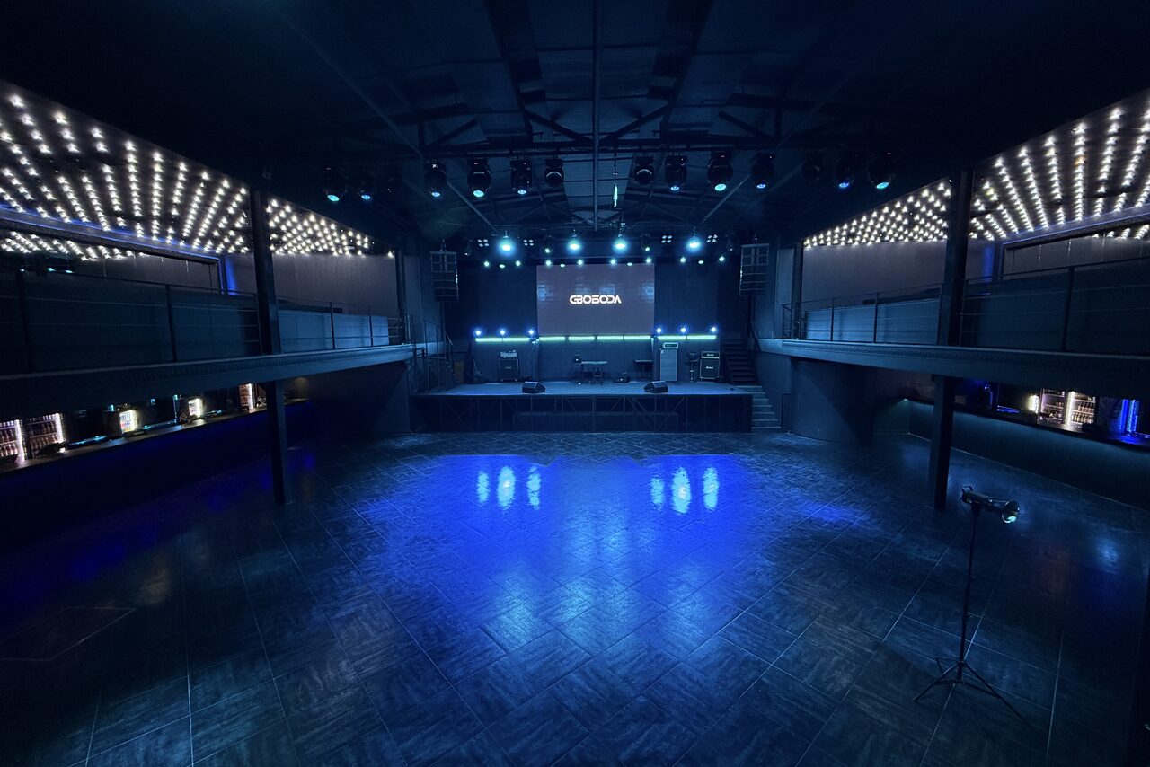 Арендовать СВОБОДА. Зал на 1200 человек. Большое помещение с сценой и танцполом, освещенное синими и белыми лампами. Пол деревянный, на сцене экран с текстом, микрофоны и динамики. По бокам возвышения с перилами. Пространство темное с акцентной подсветкой.