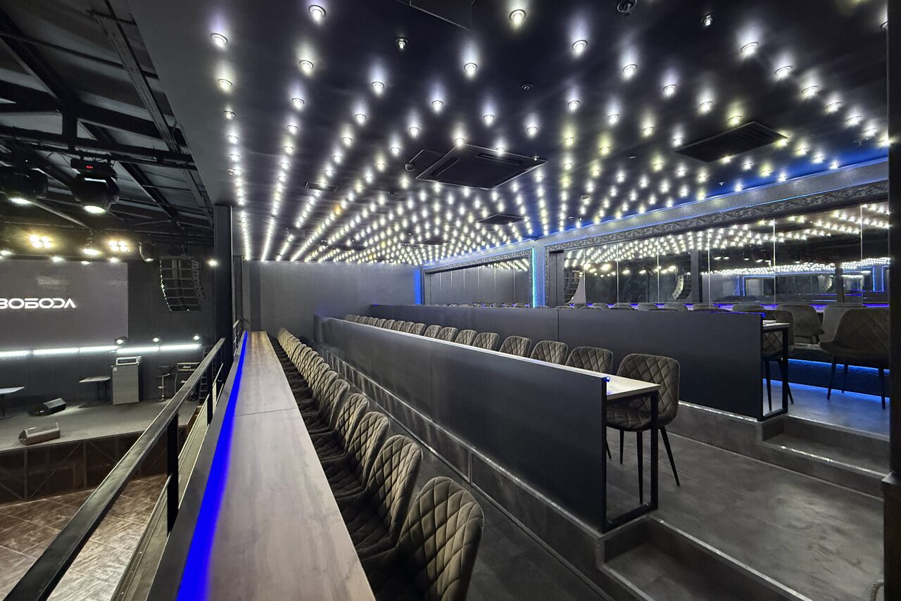 Арендовать СВОБОДА. Зал на 1200 человек. Зал с амфитеатровой рассадкой и барной стойкой, стулья с мягкой обивкой, потолок с множеством точечных светильников, серые стены, большие зеркала. Светодиодная подсветка синего цвета создает современный дизайн.