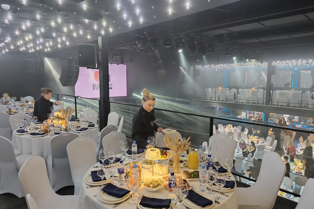 Арендовать СВОБОДА. Зал на 1200 человек. Мероприятие в банкетном зале с круглыми столами, накрытыми белыми скатертями и синими салфетками. Стулья белого цвета. Нежное освещение, свисающие лампочки. На заднем плане сцена с экраном. Люди расставляют столовые приборы, бокалы и декор.