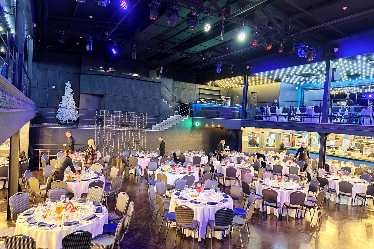 Арендовать СВОБОДА. Зал на 1200 человек. Большое помещение в стиле лофт с высоким потолком и балконом. Расставлены круглые столы с белыми скатертями и сервировкой. Присутствуют разноцветные светильники, новогодняя елка и светодиодные украшения.