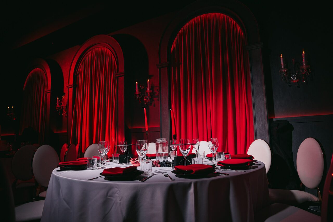 Арендовать QUQLA. Круглый стол с белой скатертью и сервировкой на шесть персон; красные салфетки, стеклянная посуда. Стулья с белой обивкой. На стенах арочные ниши с закрытыми красными шторами, красное освещение, декоративные настенные канделябры.