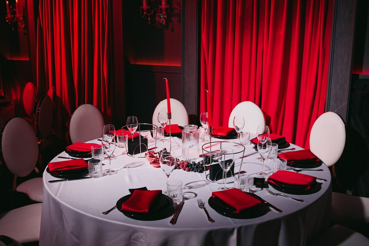 Арендовать QUQLA. Круглый обеденный стол с белой скатертью и красными салфетками, окруженный белыми стульями. На столе бокалы, тарелки, столовые приборы и свечи. Задник оформлен красными шторами. Освещение мягкое, акцент на красные и белые цвета.