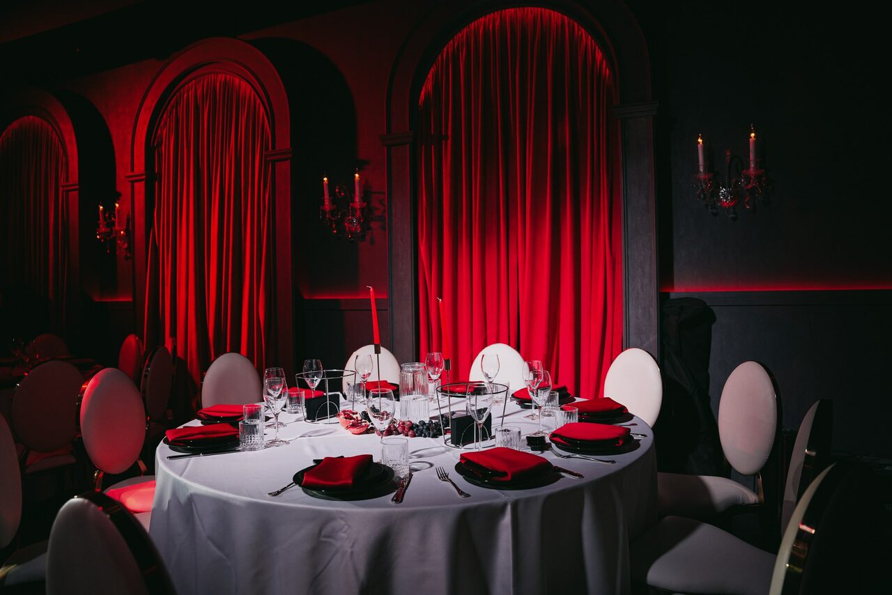 Арендовать QUQLA. Круглый стол, сервированный бокалами и столовыми приборами, окружен мягкими стульями. На столе черные тарелки с красными салфетками. Красные шторы на фоне, тёмные стены с подсвечниками. Освещение мягкое, красно-тёмная цветовая гамма.