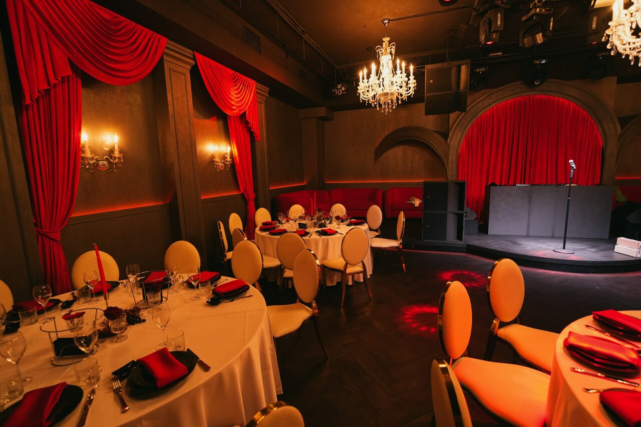 Арендовать QUQLA. Банкетный зал с круглыми столами, накрытыми белыми скатертями, сервированными бокалами и черно-красными салфетками. Стулья с кремовыми спинками. В центре сцена с микрофоном. Красные шторы, темно-коричневые стены, хрустальные люстры, мягкое освещение.