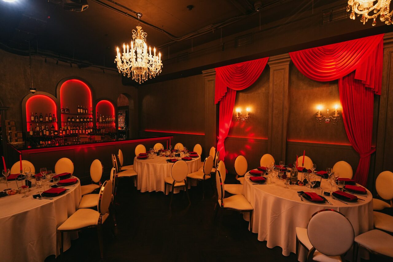 Арендовать QUQLA. Помещение для мероприятий с круглыми столами и белыми стульями. Стены украшены красными шторами и подсветкой. Пара люстр и настенные светильники обеспечивают освещение. Имеется барная стойка с полками для бутылок. Стены темно-серого цвета.