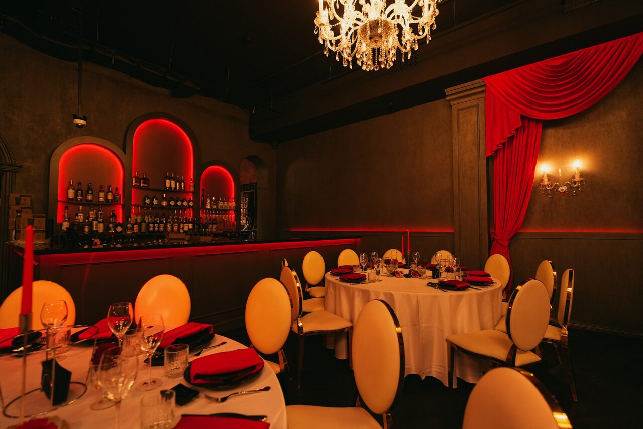 Арендовать QUQLA. Ресторанное пространство с круглыми столами, белыми скатертями, красными салфетками, бокалами и столовыми приборами. Стены с нишами для бутылок, подсвеченные красным светом. Люстра сверху, красные портьеры. Интерьер в темных тонах.