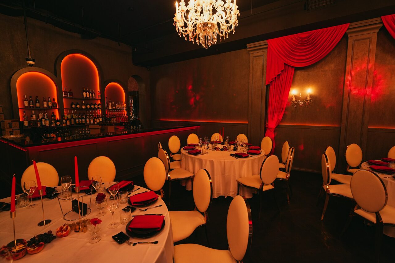 Арендовать QUQLA. Зал с барной стойкой и круглым столом, накрытым белой скатертью. Белые стулья с овальной спинкой, красные салфетки и свечи. На стенах декоративные лепные элементы, красные драпировки. Люстра и бра с мягким светом. Тёмные стены и полы.