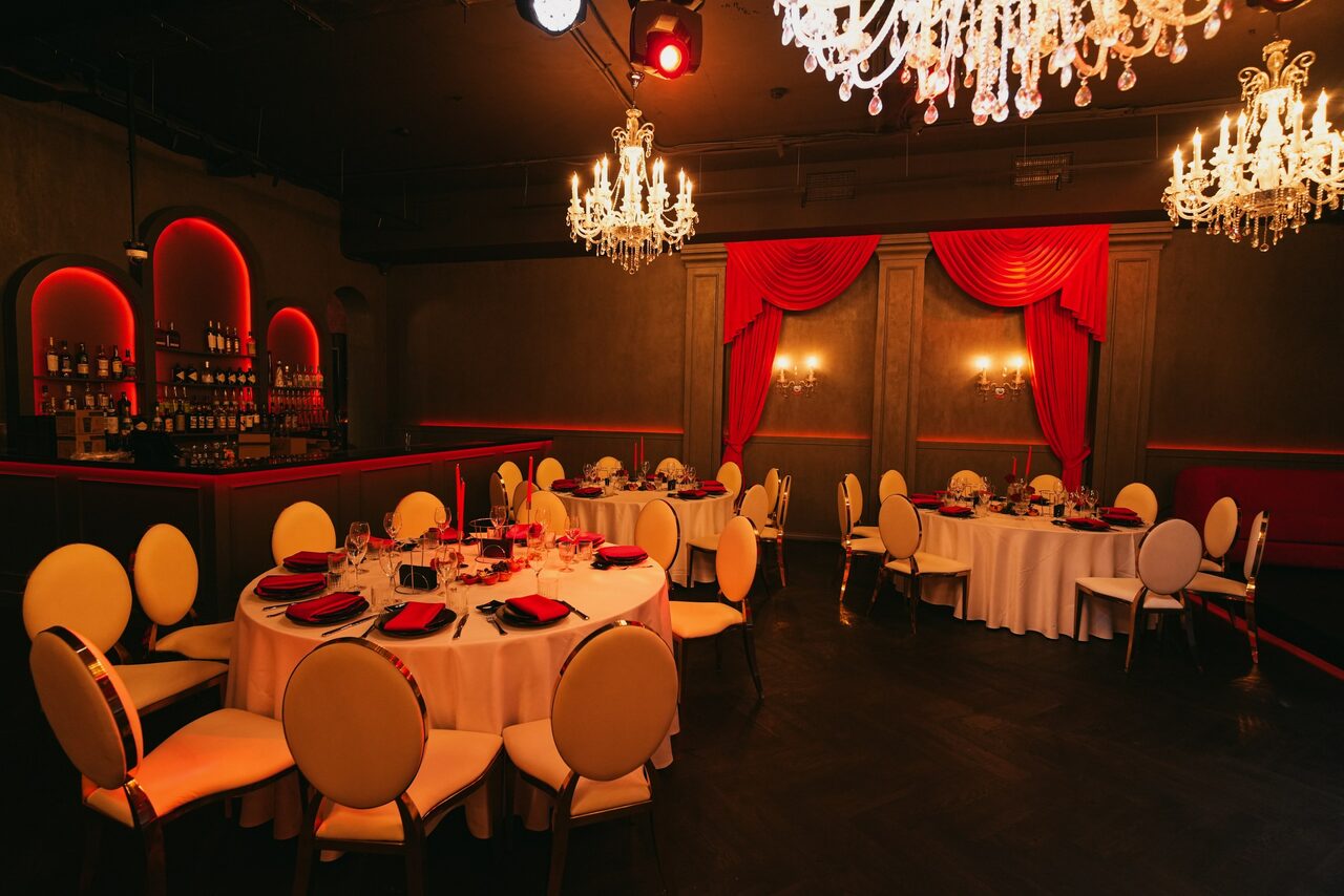Арендовать QUQLA. Банкетный зал с круглыми столами, накрытыми белыми скатертями и черными салфетками. Белые стулья с подушками. Красные драпировки на стенах. Люстра с хрустальными элементами. Бар в углу с подсветкой. Теплое освещение. Тёмные полы.