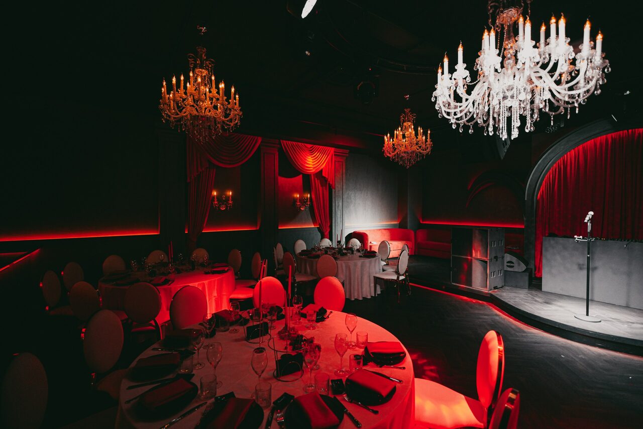 Арендовать QUQLA. Торжественное пространство с круглым столом, сервированным для мероприятия, в окружении белых стульев. Декор с красными занавесями, черными стенами и подвешенными люстрами. На сцене микрофон, зонированное освещение с теплым красным и желтым светом.