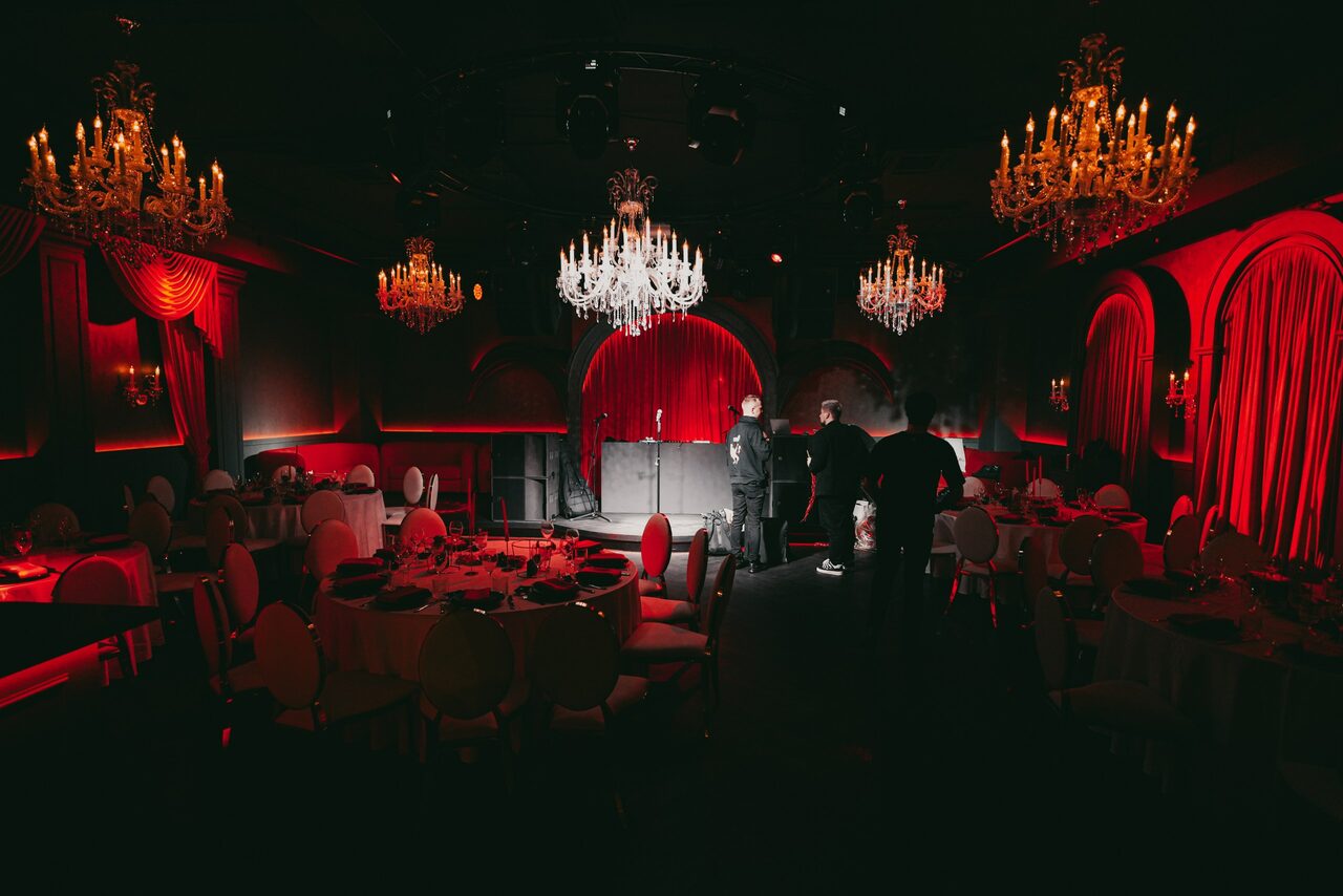 Арендовать QUQLA. Банкетный зал с несколькими круглыми столами, накрытыми на мероприятие, окружёнными стульями. Декор в красно-чёрных тонах, красные занавески, подвесные хрустальные люстры создают тёплое освещение. Люди стоят у сцены с красным задником.