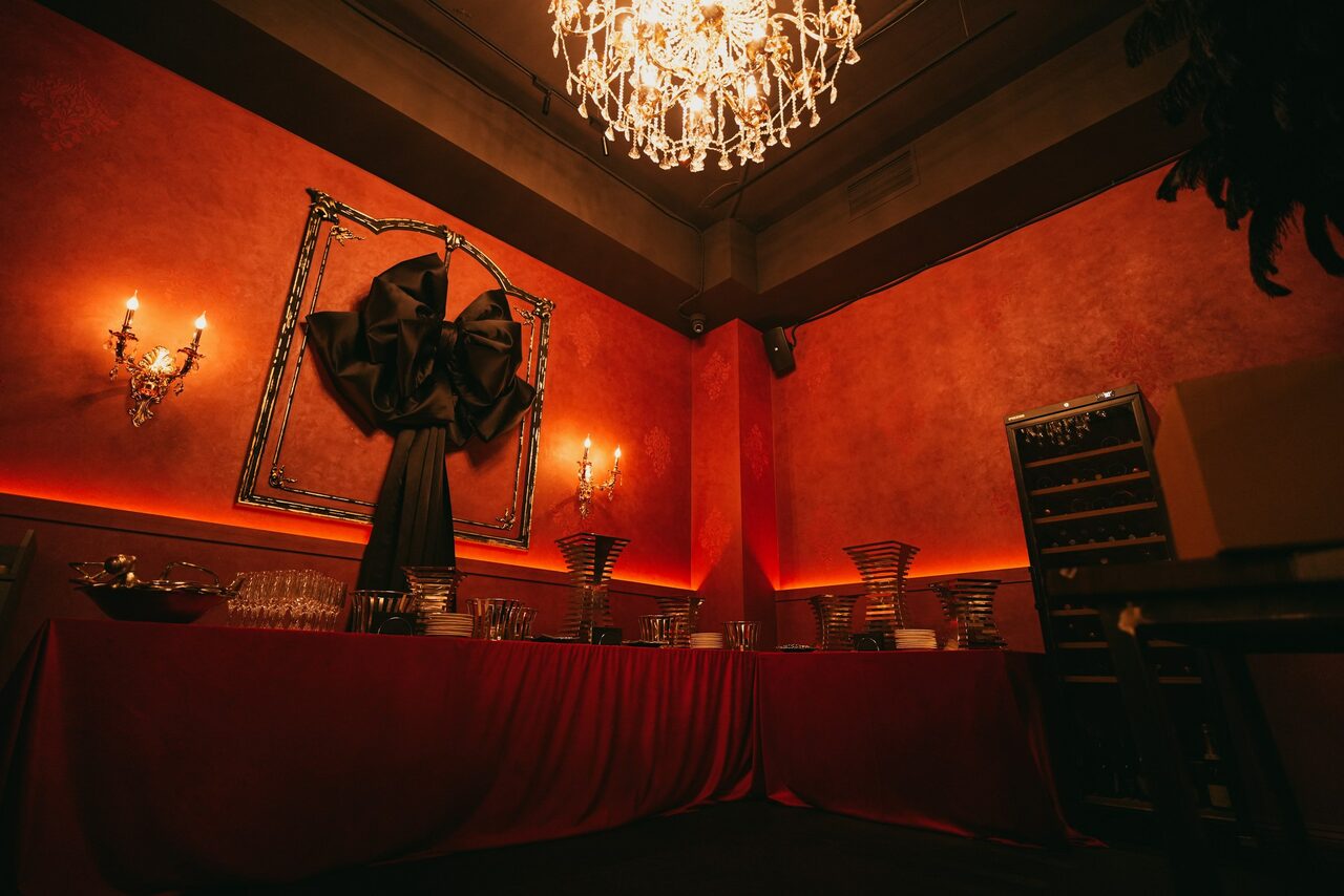 Арендовать QUQLA. Красная комната с вечерним освещением. Потолок украшает большая хрустальная люстра. Стены декорированы зеркалом с черной тканью. По периметру длинный стол с красной скатертью, посудой и подсвечниками. Справа высокий стеллаж с полками. На стенах канделябры.