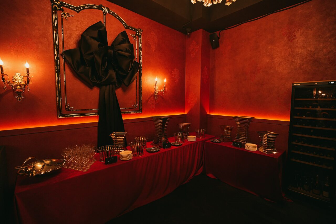 Арендовать QUQLA. Тёмное помещение с красными стенами, украшенными двумя настенными светильниками. Длинный стол вдоль стены, накрытый красной скатертью, сервирован посудой и декоративными элементами. На стене крупный чёрный бант в рамке. Справа шкаф с бокалами.