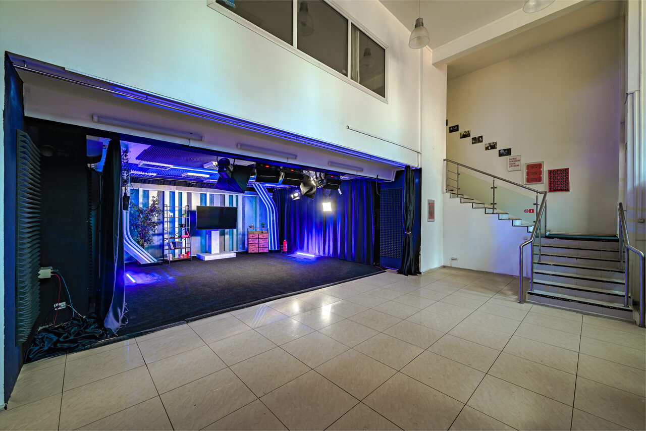 Арендовать Статус Холл. Многофункциональный зал с оборудованием. Просторное помещение с белыми стенами и плиточным полом. Сцена с черными занавесами, лампами и кабелями. Стены оформлены синим освещением. Металлическая лестница с перилами справа. Над сценой большие окна и светильники на потолке.