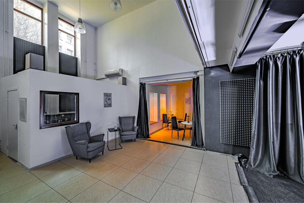 Арендовать Статус Холл. Многофункциональный зал с оборудованием. Современное многоуровневое помещение с высокими потолками. В левой части два серых кресла и столик на плиточном полу. Справа обеденная зона с круглыми столами, освещенная мягким светом. Большие окна, текстильные шторы и панели на стенах.