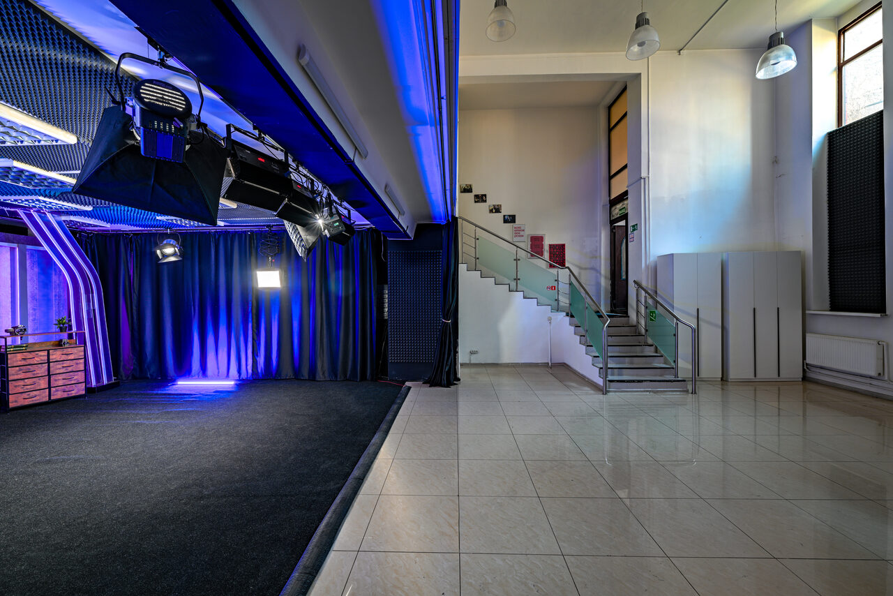 Арендовать Статус Холл. Многофункциональный зал с оборудованием. Студийное пространство с черным фоном и синим освещением, подвесное оборудование. Слева деревянный стол, справа лестница с прозрачными перилами из стекла. Пол из светлой плитки, высокие потолки, подвесные светильники.