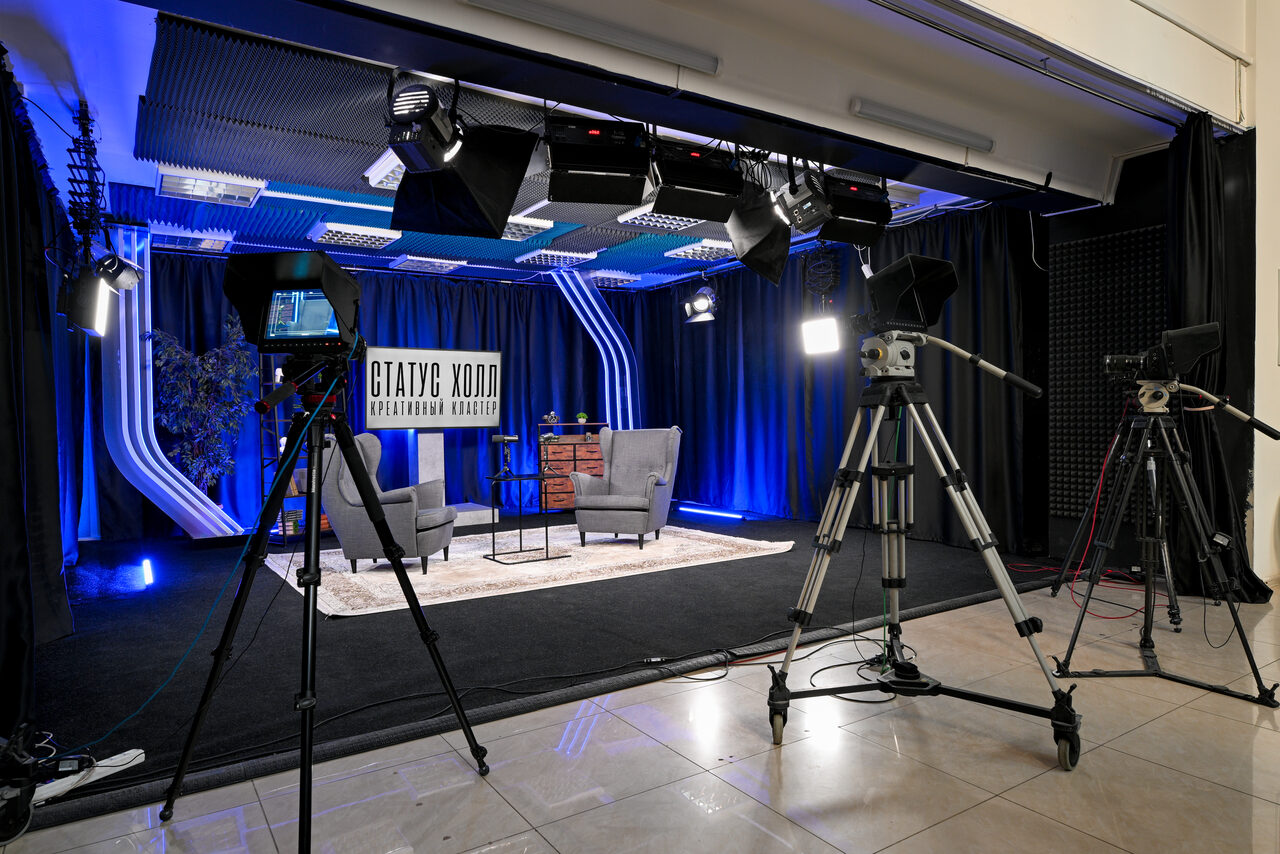 Арендовать Статус Холл. Многофункциональный зал с оборудованием. Студийное пространство с черными и синими шторами, тремя видеокамерами на штативах, серыми креслами и столом на ковре. Освещение представлено студийными светильниками. Есть знак с текстом "СТАТУС ХОЛЛ". Пол — плитка, стены — штора.