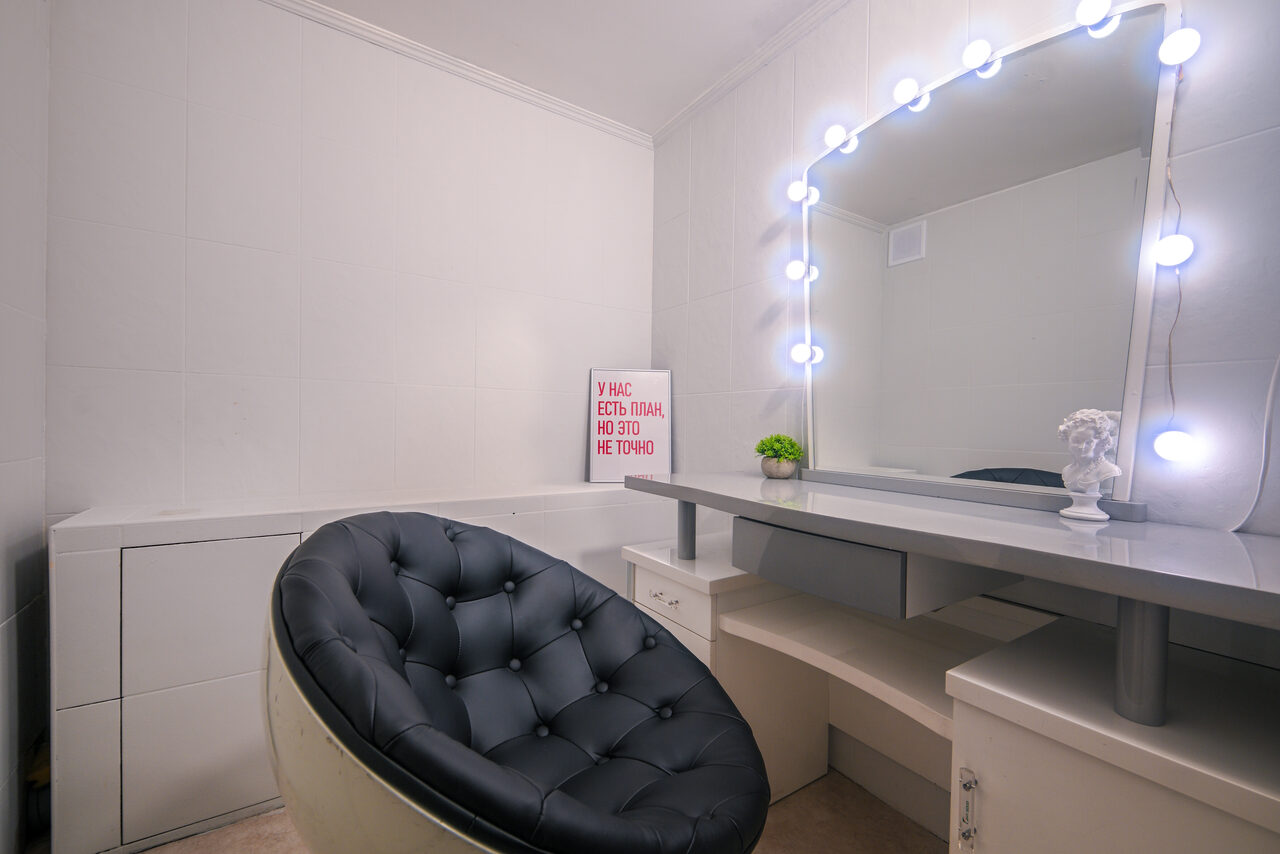 Арендовать Статус Холл. Многофункциональный зал с оборудованием. Комната с белыми стенами, туалетным столиком и круглым зеркалом с лампочками по периметру. Черное кресло с пуговицами, стол серого цвета, на нем белая статуэтка и растение. На стене постер с текстом «У нас есть план, но это не точно».