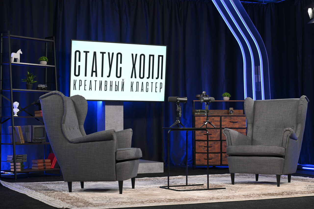 Арендовать Статус Холл. Многофункциональный зал с оборудованием. Современное студийное пространство с двумя серыми креслами, ковром и столиком на фоне темных занавесей. Освещение представлено настольными лампами. За креслами виден экран с текстом, слева - стеллаж с декором.