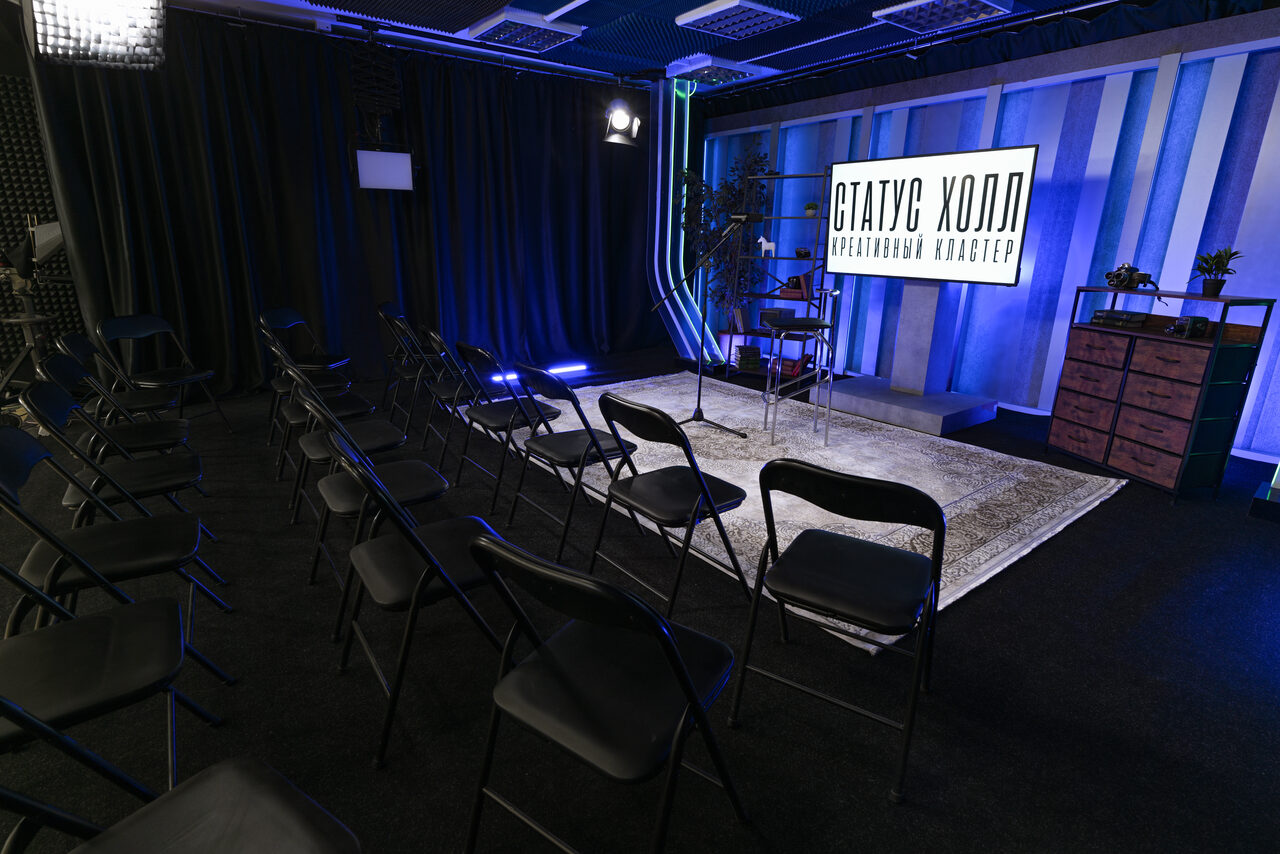 Арендовать Статус Холл. Многофункциональный зал с оборудованием. Зал с черными складными стульями в несколько рядов. На стене светодиодный экран с надписью "Статус Холл". Стены декорированы вертикальными полосами, освещение синего оттенка. Напольное покрытие темное, в центре ковер. Сбоку светильники и стойка с растениями.