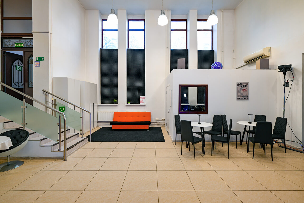 Арендовать Статус Холл. Многофункциональный зал с оборудованием. Просторное помещение с высокими потолками, деталь лестницы слева, три больших окна, черные жалюзи, оранжевый диван на черном ковре, три круглых стола с черными стульями, светлое кафельное покрытие, подвесные светильники, кондиционер, настенные знаки.