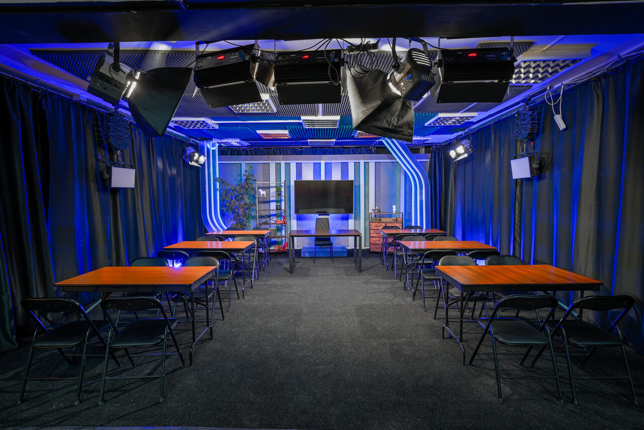 Арендовать Статус Холл. Многофункциональный зал с оборудованием. Комната с темным ковровым покрытием, расположены пять столов с деревянными крышками и складными стульями. Настенное освещение из синей светодиодной подсветки. На стене телевизор. Потолочные софиты и черные шторы вдоль стен.