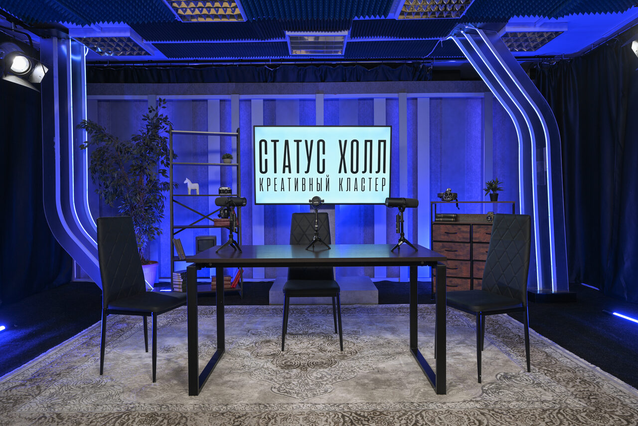 Арендовать Статус Холл. Многофункциональный зал с оборудованием. Студия с тремя стульями и черным столом на ковре, два микрофона. На заднем плане светло-синяя стена, экран с надписью «СТАТУС ХОЛЛ КРЕАТИВНЫЙ КЛАСТЕР», полки с декором, деревянный комод. Сцена освещена различными источниками света.