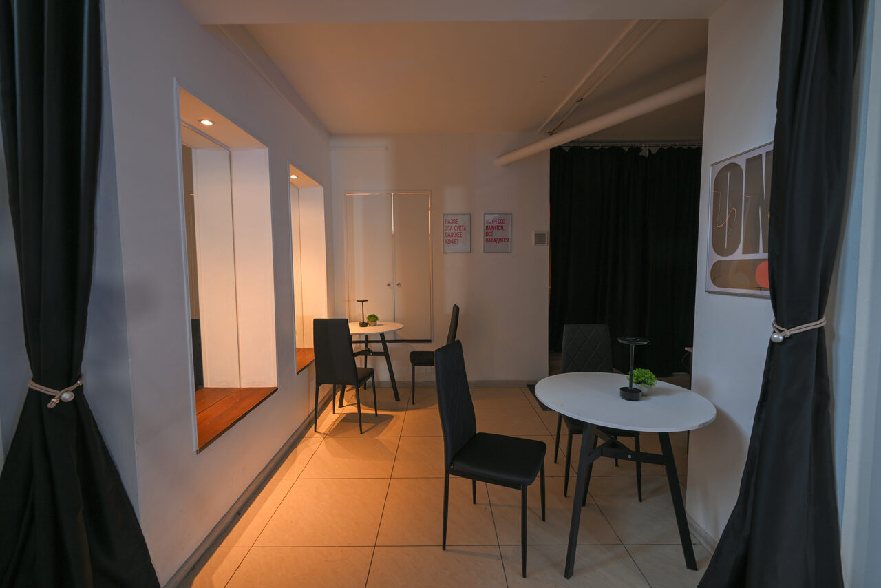 Арендовать Статус Холл. Многофункциональный зал с оборудованием. Небольшое помещение с двумя столами и черными стульями, светлая плитка на полу, стены белые с постерами и черными занавесками, встроенные ниши с зеркалами и точечными светильниками.