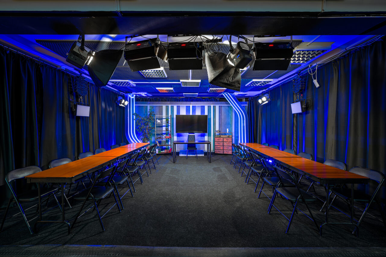 Арендовать Статус Холл. Многофункциональный зал с оборудованием. Конференц-зал с двумя длинными рядами деревянных столов и черными стульями. На стенах висят светильники. На потолке подвесные панели и студийное освещение. В центре большой экран, вокруг темные шторы. Освещение синим и белым светом.