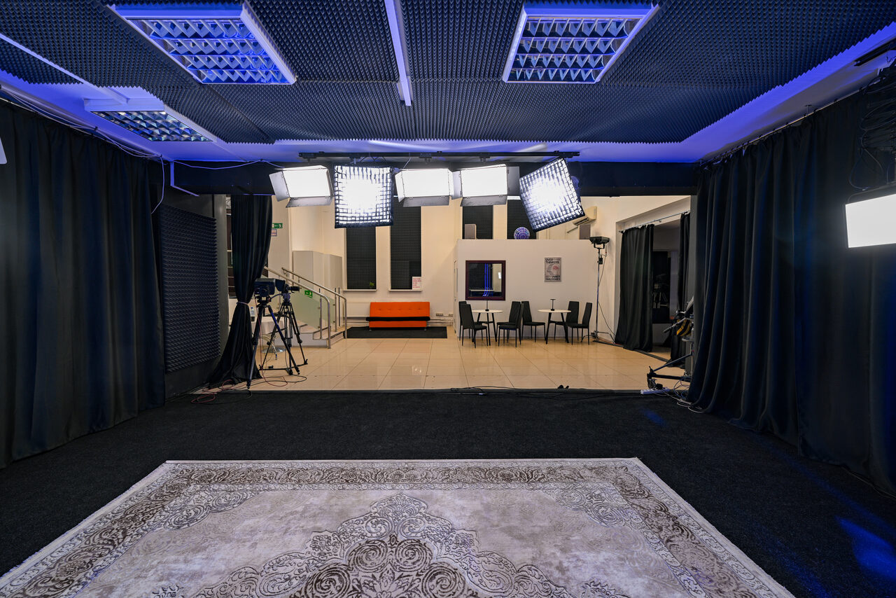 Арендовать Статус Холл. Многофункциональный зал с оборудованием. Студия с черными занавесками по периметру, потолок с осветительными приборами, на полу узорный ковер, по центру стулья и оранжевый диван. На стенах фотографии, установлены камеры и стойки для освещения.