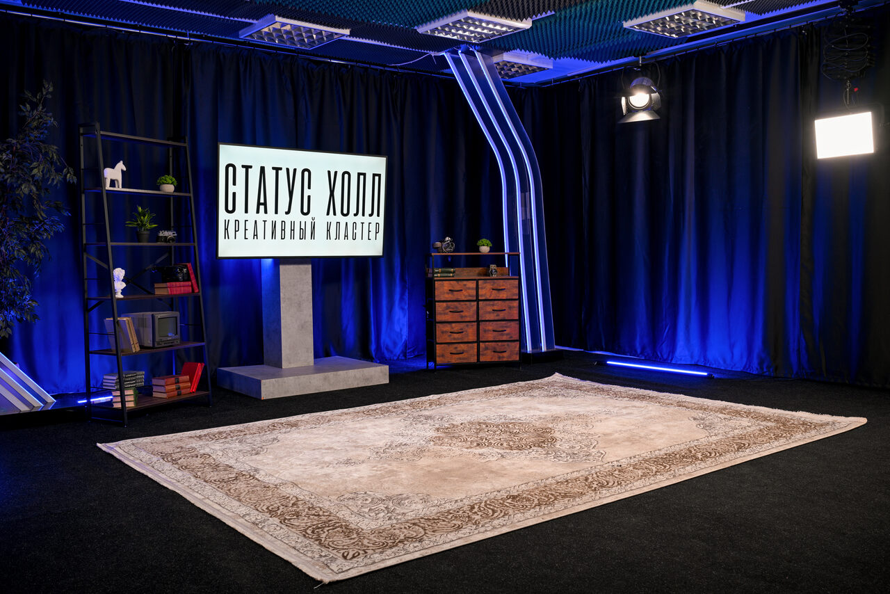Арендовать Статус Холл. Многофункциональный зал с оборудованием. Пространство с черными шторами и синим акцентным освещением. На полу бежевый ковер с узором. Металлический стеллаж с декором, экран на подиуме с текстом «СТАТУС ХОЛЛ КРЕАТИВНЫЙ КЛАСТЕР», деревянный комод, потолочные светильники.