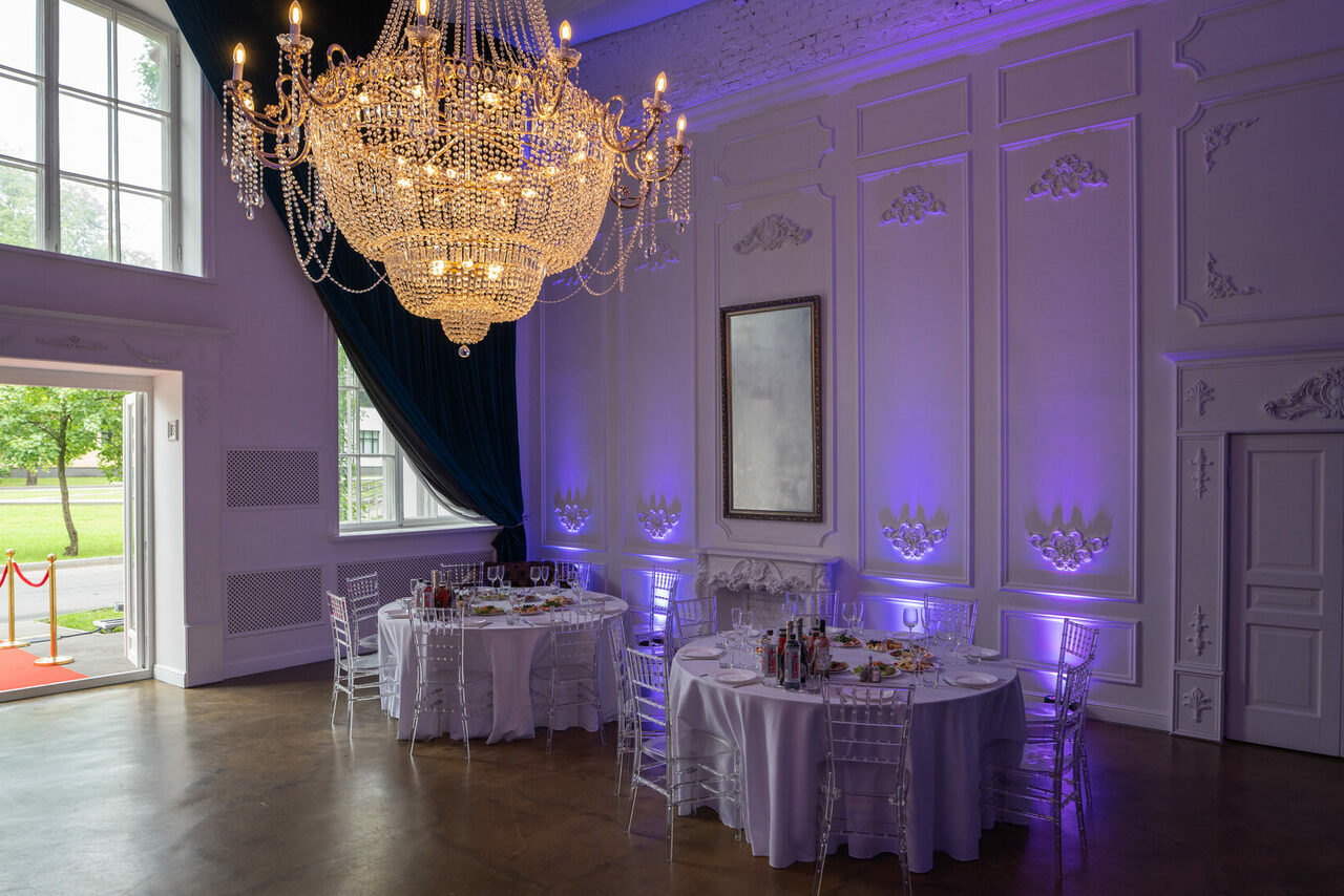 Арендовать Лофт КОСМОС с летней верандой. Просторная комната с высокими потолками; большая люстра; две круглые сервированные столы с прозрачными стульями; стены с декоративной лепниной и подсветкой фиолетового оттенка; большие окна с зелеными шторами; деревянный пол; белые двери.