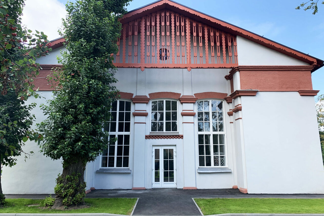 Арендовать Лофт КОСМОС с летней верандой. Фасад здания с большими арочными окнами, белыми стенами и красными декоративными элементами. Центральная дверь окружена двумя высокими окнами, крыша остроконечная. Перед зданием растет дерево, есть дорожка и зеленая лужайка.