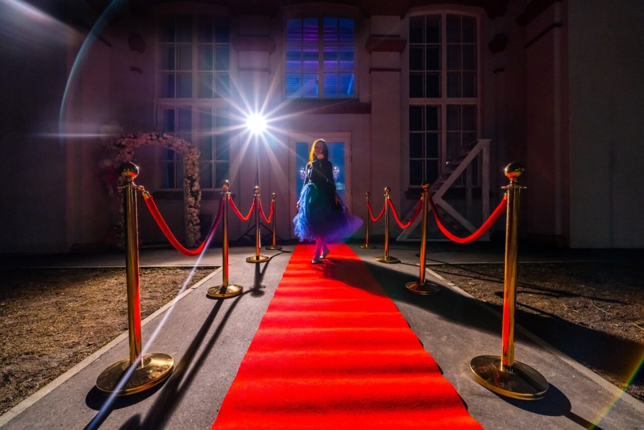 Арендовать Лофт КОСМОС с летней верандой. Красная ковровая дорожка, окруженная золотыми стойками с натянутыми красными канатами, ведет к зданию с большими окнами. Яркий прожектор освещает дорожку. Стены светлые, в углу арка украшена цветами.