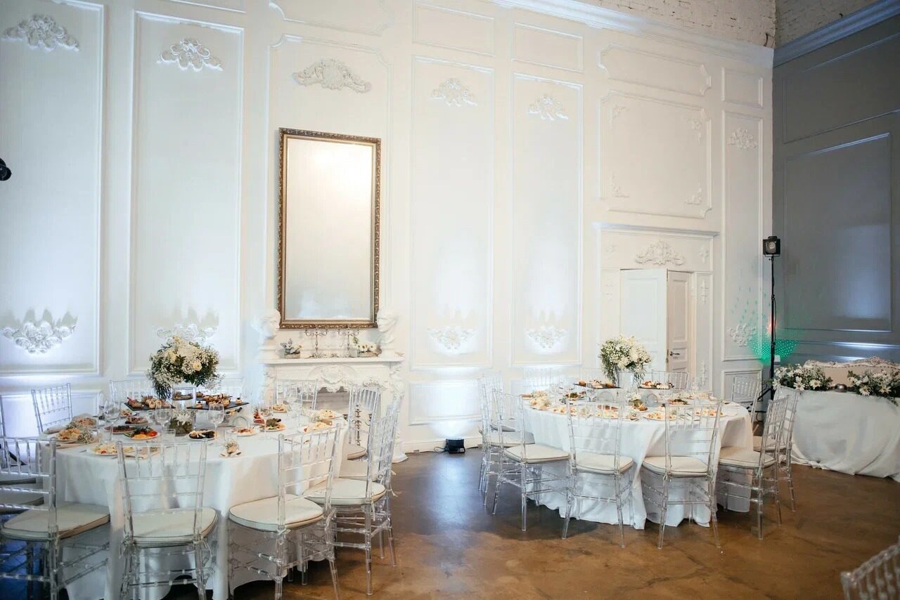 Арендовать Лофт КОСМОС с летней верандой. Белый зал с высокими панелями и декором. Круглые столы накрыты белыми скатертями, украшены цветами, окружены прозрачными стульями. На стене большое зеркало в раме, мягкое освещение. Пол светло-коричневый.