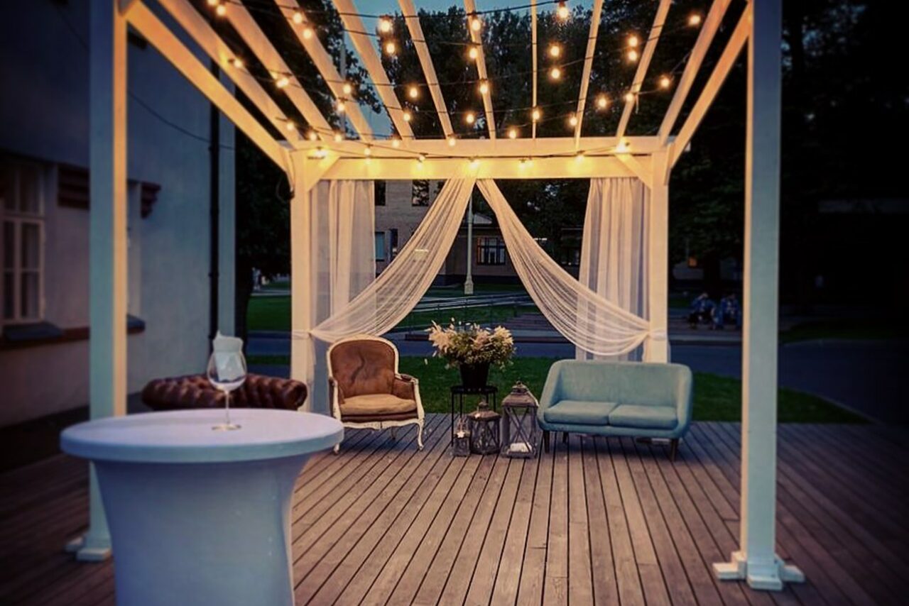 Арендовать Лофт КОСМОС с летней верандой. Пергола с подвесными огнями и тканевыми занавесками, деревянный настил. Внутри стол, кресла и диван. Светлый текстиль, декоративные цветы в вазе, фонари на полу. Декор в теплых тонах, уютная вечерняя атмосфера.