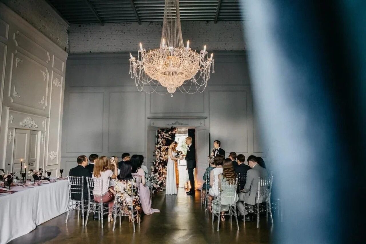 Арендовать Лофт КОСМОС с летней верандой. Просторное помещение с высокими потолками, большая люстра, бежевые стены с декоративными элементами, деревянный пол. Ряды прозрачных стульев, парализованный стол с белой скатертью слева. Декоративная арка с цветами. Собрание людей.