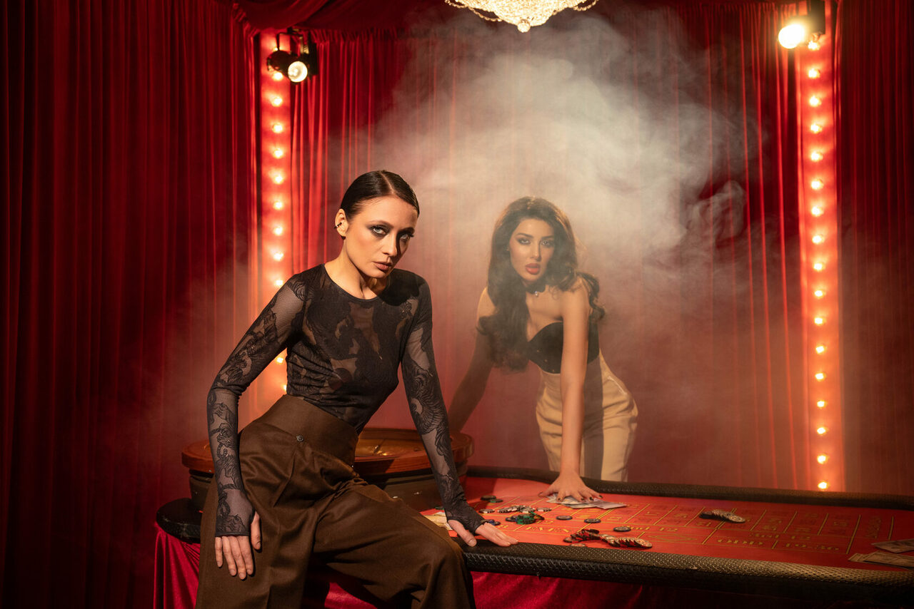 Арендовать Лофт КОСМОС с летней верандой. Красные бархатные шторы, у стены стол для игры в рулетку с фишками и картами, освещение лампочками по периметру, дымка в воздухе, две женщины у стола в вечерних нарядах.