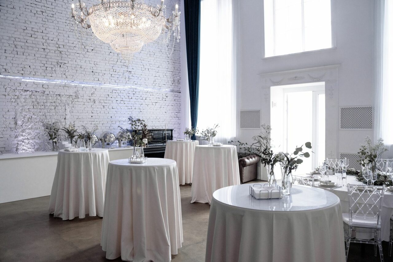 Арендовать Лофт КОСМОС с летней верандой. Зал с высоким потолком и белыми кирпичными стенами. Круглые столы с белыми скатертями, украшенные цветами. Большая люстра, диван, стулья, высокие окна с занавесками. Светлые тона, натуральное освещение, двери с резными элементами.