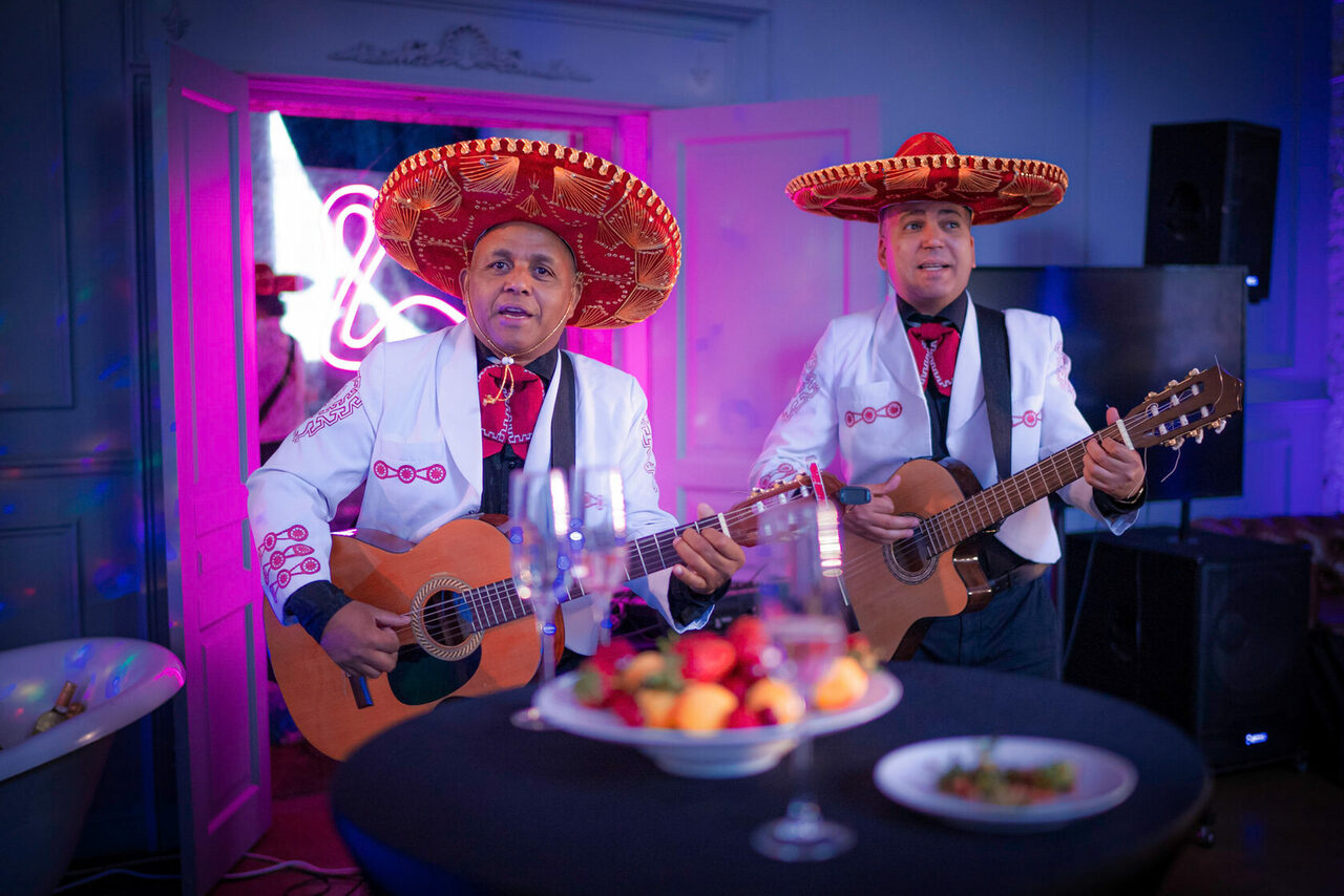 Арендовать Лофт КОСМОС с летней верандой. Музыкальная сцена, два мужчины в белых костюмах и сомбреро играют на гитарах. Перед ними высокий стол с фруктами и бокалами. На стене неоновый знак. Темные стены, яркое разноцветное освещение.