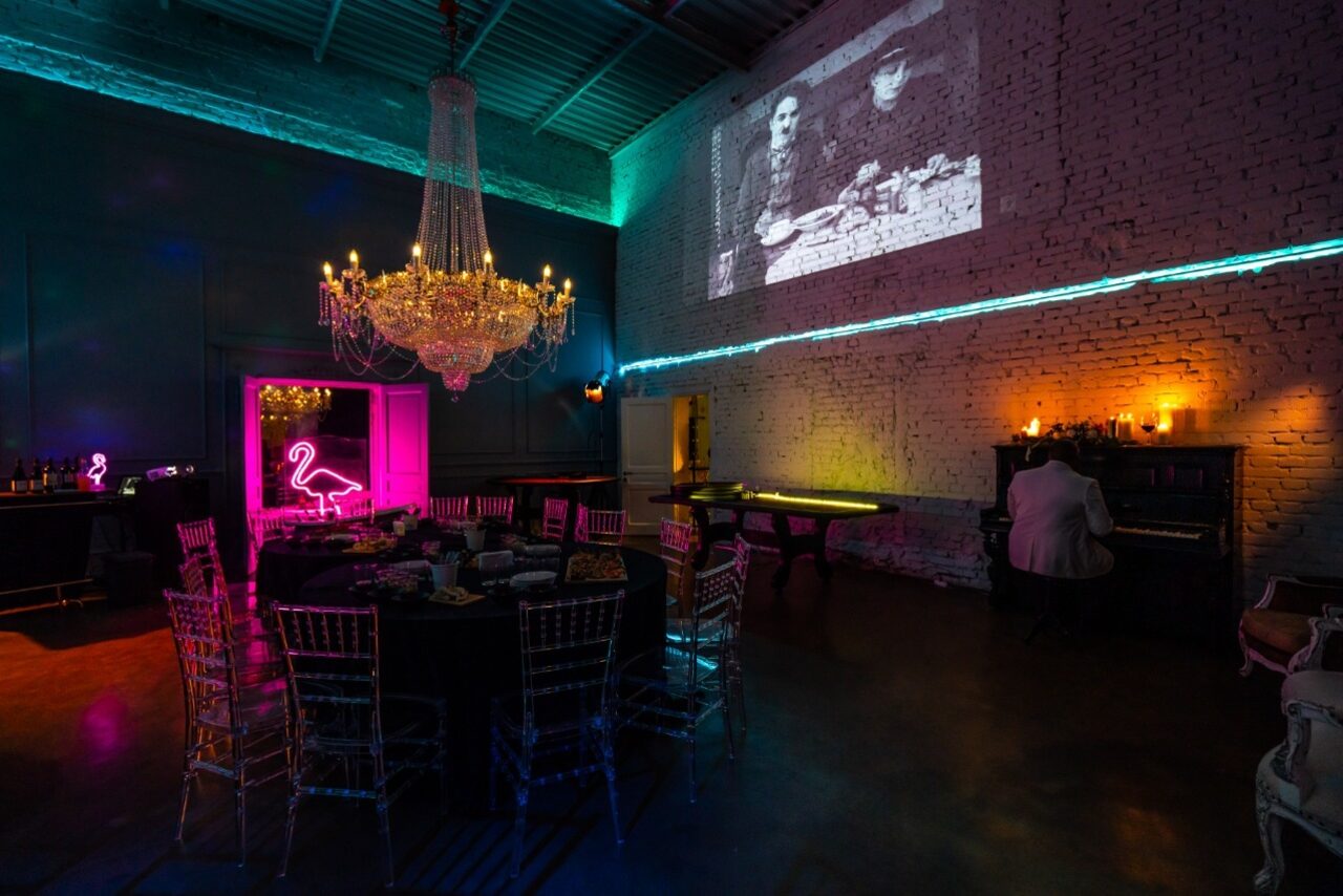 Арендовать Лофт КОСМОС с летней верандой. Большое помещение с кирпичными стенами, деревянными полами и высоким потолком. Центральная часть занята круглым столом и стульями. На стене висят неоновые вывески, большая люстра, проектор показывает изображение. Атмосферное освещение.