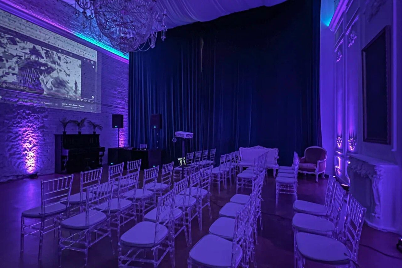 Арендовать Лофт КОСМОС с летней верандой. Зал с рядами прозрачных стульев, направленных на большую проекцию на стене. Слева от экрана — музыкальная аппаратура, освещение фиолетовое и голубое. Потолок украшен люстрой, стены из белого кирпича и декорированы лепниной.