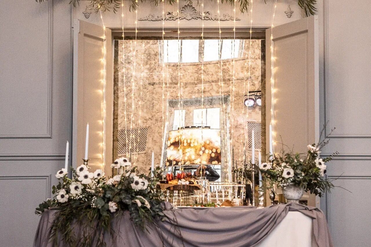 Арендовать Лофт КОСМОС с летней верандой. Светло-серое помещение с большим окном, украшенное световыми гирляндами и цветочной композицией. На переднем плане стол с серой тканью, свечами и цветами. На заднем плане зеркало и светильники.