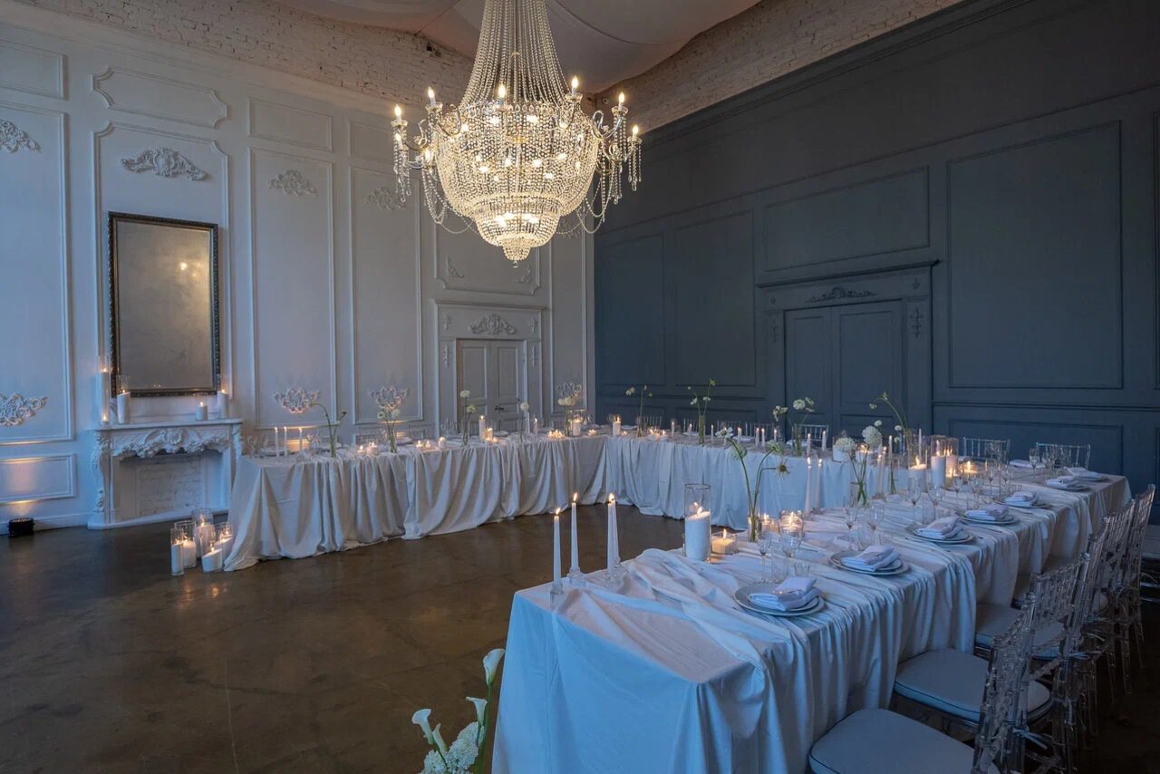 Арендовать Лофт КОСМОС с летней верандой. Зал с длинными сервированными столами с белыми скатертями, стулья из прозрачного пластика. На столах свечи и бокалы, освещение от большого подвесного люстра. Стены белого и серого цвета, декоративные панели, высокое зеркало на стене. Пол темного цвета.