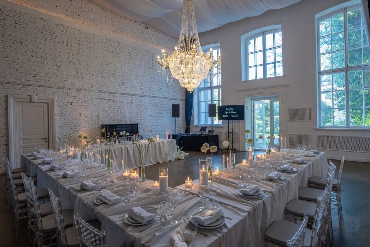 Арендовать Лофт КОСМОС с летней верандой. Большой зал с кирпичными стенами, высокой потолочной люстрой и большими окнами. Расставлены обеденные столы в П-образной форме с белыми скатертями, прозрачные стулья, свечи и сервировка. На заднем плане колонка и экран, дверь и вид на улицу.