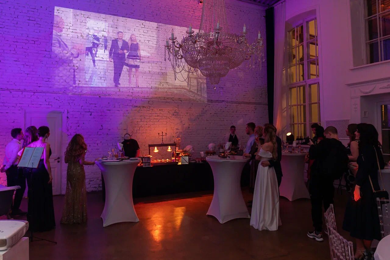 Арендовать Лофт КОСМОС с летней верандой. Пространство для мероприятий с высоким потолком, проекцией на кирпичной стене, большой люстрой. Белые барные столы, гости в вечерних нарядах. Декор в тёплых тонах, деревянный пол. Окна с белыми рамами. Освещение разноцветное.