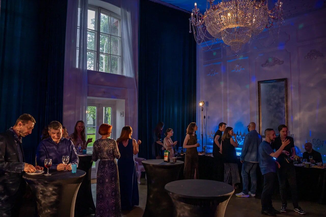 Арендовать Лофт КОСМОС с летней верандой. Просторное помещение с высокими потолками, люстра, большие окна с синими занавесками. Расставлены круглые высокие столы без скатертей. Люди общаются, игры света и тени на стенах. На стене большое зеркало.