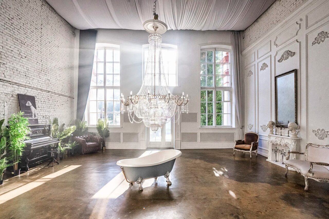 Арендовать Лофт КОСМОС с летней верандой. Просторное помещение с высокими потолками и большими окнами. На потолке подвешена большая люстра, по центру стоит ванна. Вдоль стен есть кресла, пианино и зелёные растения. Стены белые с декоративными элементами, пол темный, светлый текстиль на потолке.