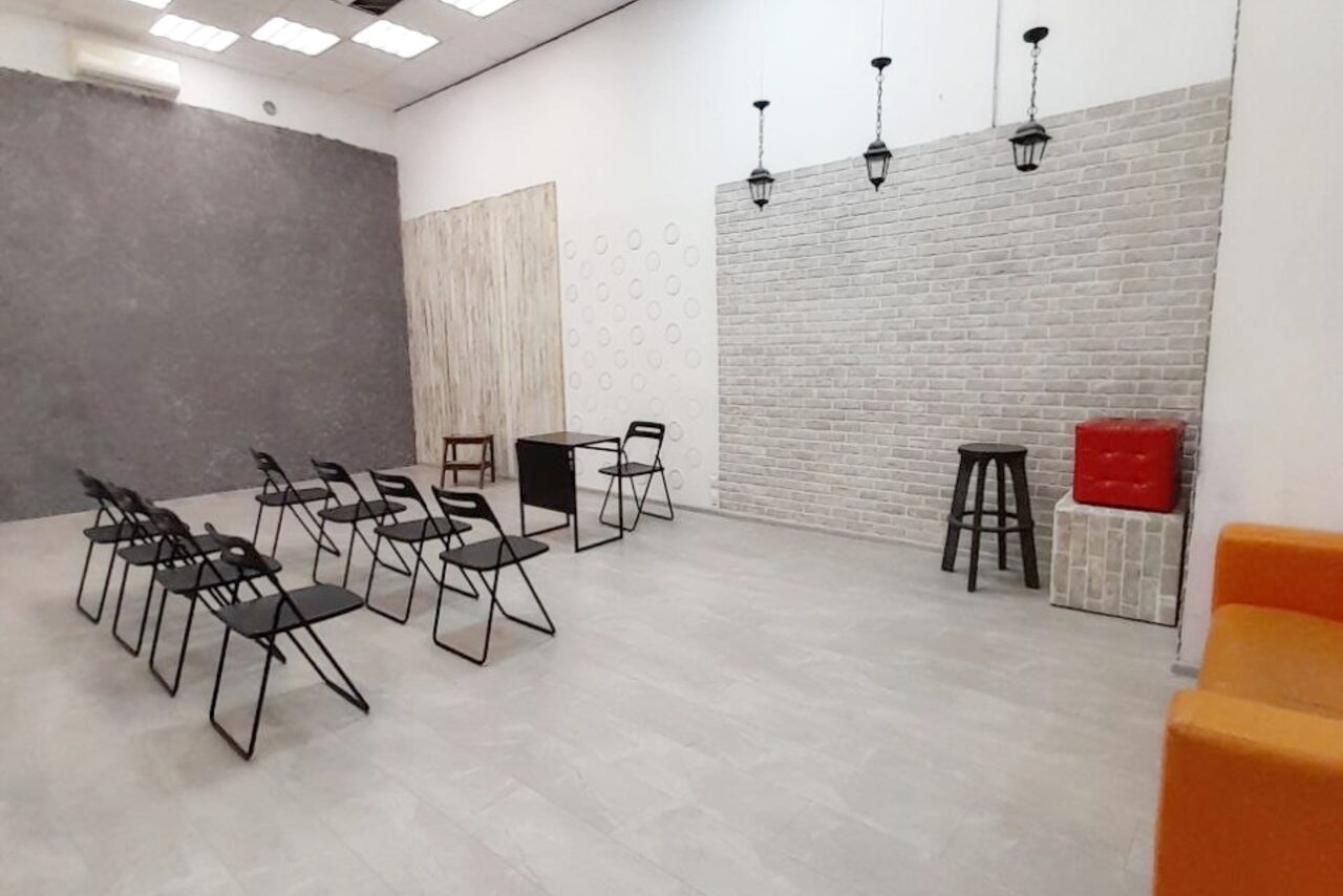 Арендовать Интер-Фото. Просторное помещение с серыми плиточными полами и белыми кирпичными стенами. Несколько рядов черных складных стульев, черный стол, высокий табурет. Ярко-оранжевое и красное сиденья. Подвесные светильники, квадратные потолочные панели.